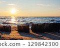 Wooden breakwater stretches into blue sea under soft evening light. Gentle waves touch sandy shore while sun reflects warmly on weathered timber posts and shimmering water 129842300