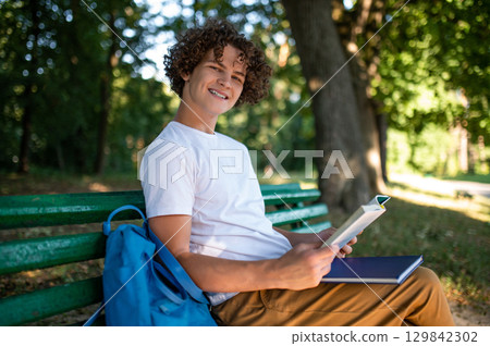 Curly-haired teenager sitting on the bench in the park and reading a book 129842302