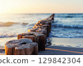 Wooden breakwater stretches into blue sea under soft evening light. Gentle waves touch sandy shore while sun reflects warmly on weathered timber posts and shimmering water 129842304