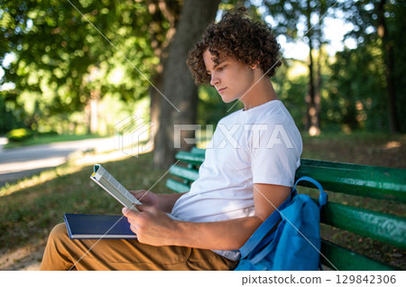 Curly-haired teenager sitting on the bench in the park and reading a book 129842306