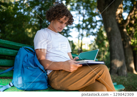 Curly-haired teenager sitting on the bench in the park and reading a book Curly-haired teenager sitting on the bench in the park and reading a book 129842309