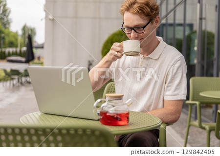 Ginger male freelancer drinking tea and checking emails using laptop 129842405