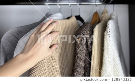 A woman sorts through clothes hanging on hangers in a closet. Reviewing and preparing seasonal wardrobe with warm knitted sweaters and jumpers for autumn winter. Minimalistic clothes organization 129843325