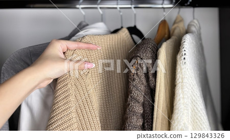 A woman sorts through clothes hanging on hangers in a closet. Reviewing and preparing seasonal wardrobe with warm knitted sweaters and jumpers for autumn winter. Minimalistic clothes organization 129843326
