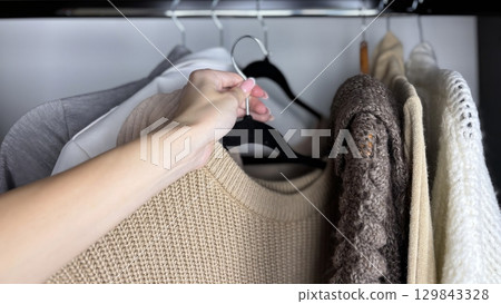 A woman sorts through clothes hanging on hangers in a closet. Reviewing and preparing seasonal wardrobe with warm knitted sweaters and jumpers for autumn winter. Minimalistic clothes organization A woman sorts through clothes hanging on hangers in a closet. Reviewing and preparing seasonal wardrobe with warm knitted sweaters and jumpers for autumn winter. Minimalistic clothes organization 129843328