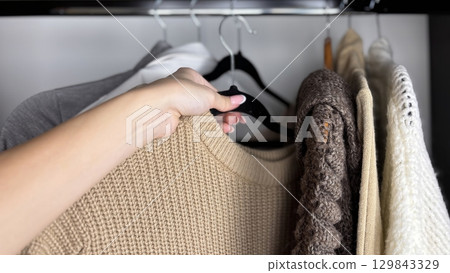 A woman sorts through clothes hanging on hangers in a closet. Reviewing and preparing seasonal wardrobe with warm knitted sweaters and jumpers for autumn winter. Minimalistic clothes organization 129843329