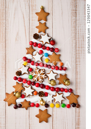Christmas tree made of gingerbread cookies, icing, candy, and baubles on wooden background, creative festive holiday composition Christmas tree made of gingerbread cookies, icing, candy, and baubles on wooden background, creative festive holiday composition 129843347