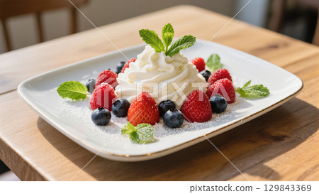 Whipped cream dessert garnished with fresh strawberries, raspberries, blueberries, and mint leaves on white plate. Light and sweet treat. 129843369