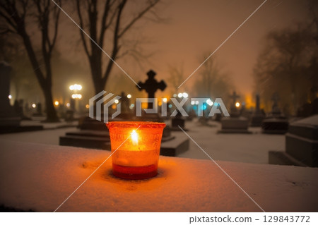 A Glowing Candle Gently Illuminates a Snowy Cemetery Throughout the Enigmatic Night Hours 129843772