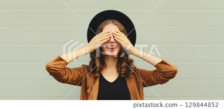 Stylish surprised young woman covering her eyes with hands, fashionable girl in black round hat 129844852