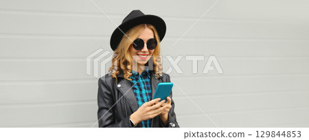 Stylish happy smiling modern young woman with phone, girl holding smartphone in black hat, jacket 129844853