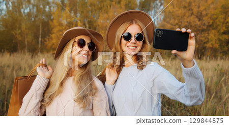 Happy women friends taking selfie with phone, shopping bags in autumn, girlfriends smiling in park Happy women friends taking selfie with phone, shopping bags in autumn, girlfriends smiling in park 129844875