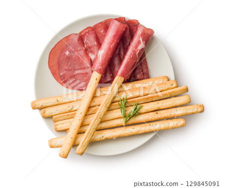 Slices of smoked bresaola and grissini sticks isolated on white background. 129845091