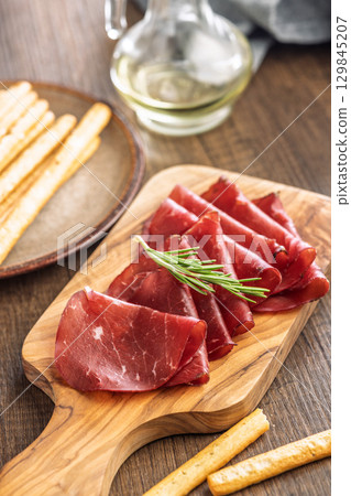 Slices of smoked bresaola on cutting board on wooden table. Slices of smoked bresaola on cutting board on wooden table. 129845207