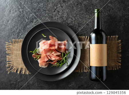 Bottle of red wine and prosciutto on a black plate. 129845356