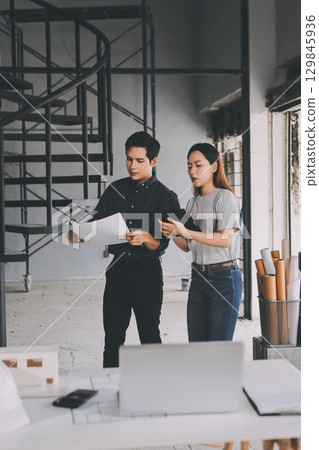 Construction manager and engineer dressed in orange work vests and hard helmets explore construction documentation on the building site near the steel frames Construction manager and engineer dressed in orange work vests and hard helmets explore construction documentation on the building site near the steel frames 129845936