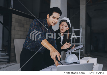 Construction manager and engineer dressed in orange work vests and hard helmets explore construction documentation on the building site near the steel frames 129845951