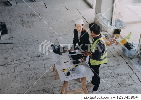 Construction manager and engineer dressed in orange work vests and hard helmets explore construction documentation on the building site near the steel frames Construction manager and engineer dressed in orange work vests and hard helmets explore construction documentation on the building site near the steel frames 129845964
