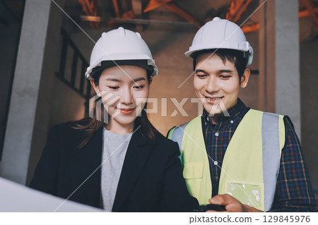 Construction manager and engineer dressed in orange work vests and hard helmets explore construction documentation on the building site near the steel frames 129845976
