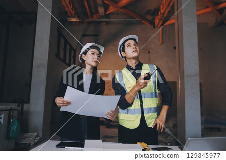 Construction manager and engineer dressed in orange work vests and hard helmets explore construction documentation on the building site near the steel frames 129845977