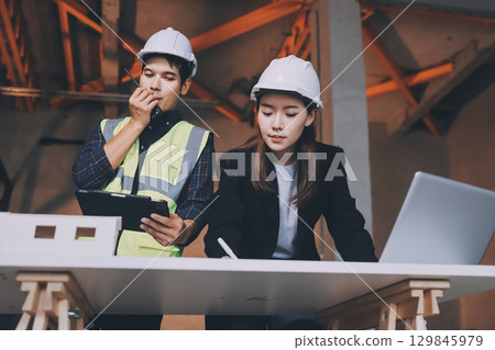 Construction manager and engineer dressed in orange work vests and hard helmets explore construction documentation on the building site near the steel frames Construction manager and engineer dressed in orange work vests and hard helmets explore construction documentation on the building site near the steel frames 129845979