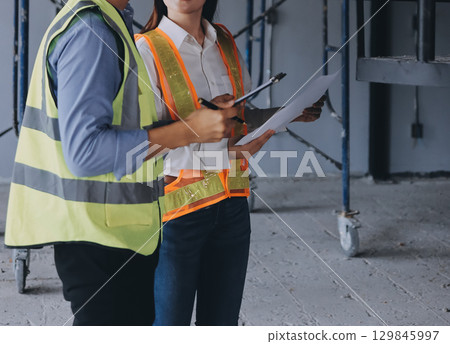 Construction manager and engineer dressed in orange work vests and hard helmets explore construction documentation on the building site near the steel frames 129845997