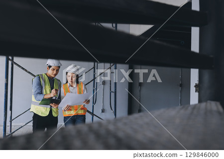 Construction manager and engineer dressed in orange work vests and hard helmets explore construction documentation on the building site near the steel frames Construction manager and engineer dressed in orange work vests and hard helmets explore construction documentation on the building site near the steel frames 129846000