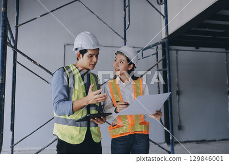 Construction manager and engineer dressed in orange work vests and hard helmets explore construction documentation on the building site near the steel frames Construction manager and engineer dressed in orange work vests and hard helmets explore construction documentation on the building site near the steel frames 129846001