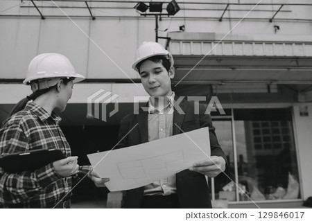 Construction manager and engineer dressed in orange work vests and hard helmets explore construction documentation on the building site near the steel frames 129846017