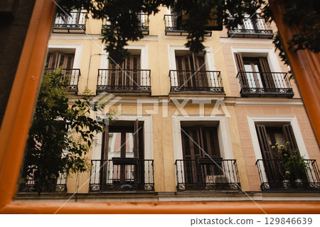 An old European building facade with windows and balconies in warm colors, bathed in sunlight. View of an old building facade featuring windows with black shutters and balconies. European city street. 129846639
