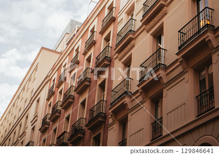 An old European building facade with windows and balconies in warm colors, bathed in sunlight. View of an old building facade featuring windows with black shutters and balconies. European city street. 129846641