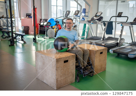Determined athlete in wheelchair do controlled lifts with weighted ball between parallel bars in gym 129847175