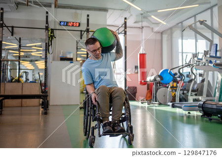 Man in wheelchair do heavy ball exercise in gym. Adaptive sports training for upper body strength Man in wheelchair do heavy ball exercise in gym. Adaptive sports training for upper body strength 129847176