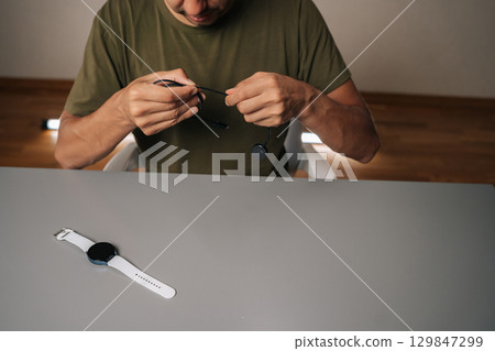 Cropped shot of focused man untangling black charging cable for smartwatch sitting at table with watch, importance of keeping device powered up. Concept of healthy lifestyle. Cropped shot of focused man untangling black charging cable for smartwatch sitting at table with watch, importance of keeping device powered up. Concept of healthy lifestyle. 129847299