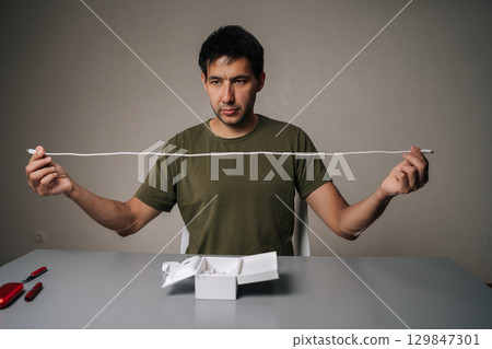 Portrait of concentrated young man unpacking new electronic cigarette at home, checking charging cable length sitting at grey table with open box. Studio shot, copy space, isolated background. Portrait of concentrated young man unpacking new electronic cigarette at home, checking charging cable length sitting at grey table with open box. Studio shot, copy space, isolated background. 129847301