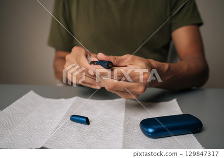 Man cleaning electronic cigarette emphasizing importance of hygiene and maintenance in vaping sitting at table with on white paper towel. Process of maintaining heated electronic cigarette device. Man cleaning electronic cigarette emphasizing importance of hygiene and maintenance in vaping sitting at table with on white paper towel. Process of maintaining heated electronic cigarette device. 129847317