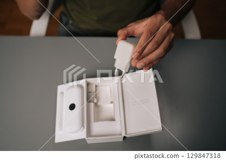 High-angle view of unrecognizable man unpacking box, holding white smartphone charger being unpacked from cardboard box, sitting on gray table. Concept of contemporary gadget unboxing. High-angle view of unrecognizable man unpacking box, holding white smartphone charger being unpacked from cardboard box, sitting on gray table. Concept of contemporary gadget unboxing. 129847318