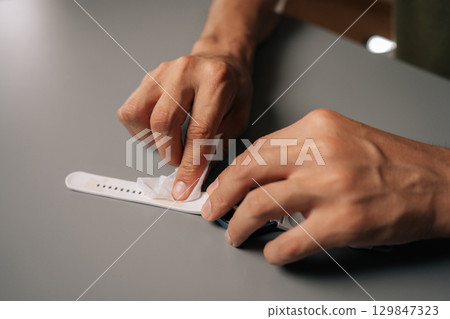 Close-up view of male hands carefully cleaning white smart watch strap with disinfectant wipe sitting at table. Concept of hygiene and proper maintenance for wearable technology. 129847323