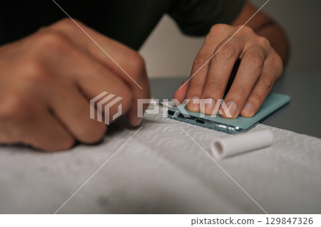 Close-up hands of repairman cleaning smartphone using specialized tools sitting at table, ensuring optimal quality and device performance. Concept of mobile phone maintenance and service. Close-up hands of repairman cleaning smartphone using specialized tools sitting at table, ensuring optimal quality and device performance. Concept of mobile phone maintenance and service. 129847326