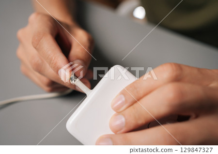 Technician hands inspecting damaged charging cable, revealing connectivity risks from visible cable fraying and potential electrical safety concerns, sitting at desk in workshop. Technician hands inspecting damaged charging cable, revealing connectivity risks from visible cable fraying and potential electrical safety concerns, sitting at desk in workshop. 129847327