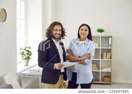 Two business people man and woman standing in office and looking cheerful at camera. Two business people man and woman standing in office and looking cheerful at camera. 129847985