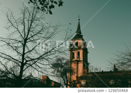 The beautiful tower of an old church stands tall on a sunny day, with autumn leaves. A historic church tower rises against a pastel blue sky, framed by tree branches and autumn leaves in silhouette. The beautiful tower of an old church stands tall on a sunny day, with autumn leaves. A historic church tower rises against a pastel blue sky, framed by tree branches and autumn leaves in silhouette. 129850468