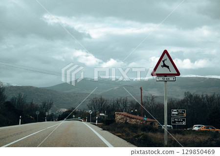 A road sign warns of deer crossing ahead and mountains under gloomy cloudy sky. The blend of nature and road travel. 129850469