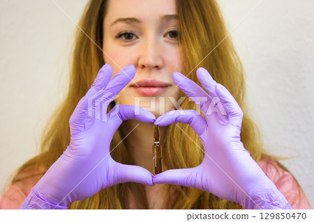 Woman holding ampoule in heart shape, symbolizing beauty and skincare. A professional beautican in purple gloves holds an ampoule in hands, making a heart shape, the concept of skincare and treatments 129850470
