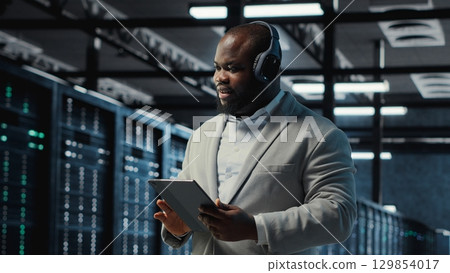 Server room programmer wearing headphones troubleshooting errors using software on tablet. IT specialist listening music, walking in data center fixing bugs using device , camera B 129854017