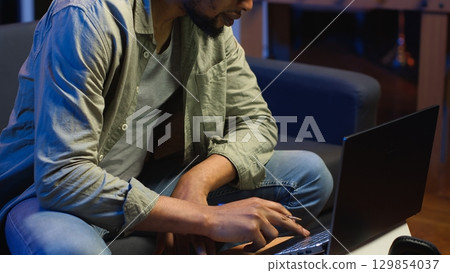 Black freelancer taking notes on textbook in order to solve his tasks, doing research and studying articles online. Young adult enjoying remote job and relaxed schedule, telework. Camera B. 129854037