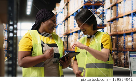Multiethnic team in hi vis vests managing retail business operations, doing product handling from the warehouse big racks. Colleagues overseeing import export activity. Camera B. 129854041