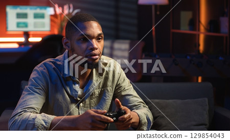 African american man spends free time playing video games with popcorn snack, having fun during game night in his cozy loft. Young adult connecting his console to the television. Camera B. 129854043