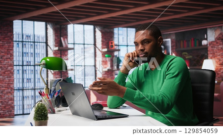 Freelancer in home office having important phone call with business partner using his mobile phone. African american guy sitting at desk discussing company matters during call. Camera A. 129854059