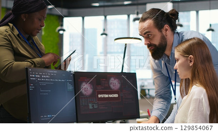 Young programmer in office requests help from IT experts detecting and blocking hacker intrusion. Intern assisted by staff members coordinating incident response for network attack, camera A 129854067